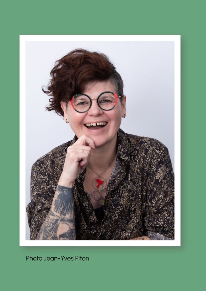 Photo de face de Béatrice Schaller-Le Leu. Buste et visage. Elle a les cheveux courts avec une mèche sur le côté et le côté gauche rasé. Elle porte des lunettes. Elle rit. Elle a une chemise marron, les manches remontées. Son bras droit est plié et soutien son menton. Elle a des tatouages apparents sur les deux bras. La photo a été prise par Jean-Yves Piton